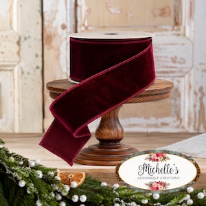 May include: A spool of deep burgundy velvet ribbon with a smooth, soft texture. The ribbon is wrapped around a central core and features a long, flowing end that drapes downward. The rich, dark red fabric has a subtle sheen across its surface.