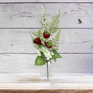 May include: A clear glass vase contains a floral arrangement with three red strawberries, white flowers, and green fern-like leaves. The arrangement is set on a white wooden surface against a white wood-panelled backdrop.