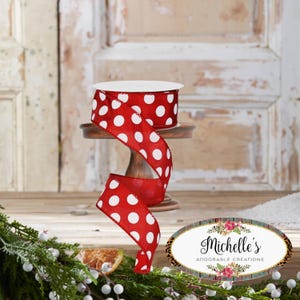 May include: A roll of red ribbon with large white polka dots sits on a small wooden stand. The ribbon is unrolled, with a portion cascading down. The background features a rustic wooden door and decorative greenery.