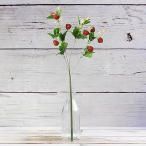 May include: A single artificial strawberry stem with red berries, green leaves, and small yellow flowers. The stem is in a clear glass vase. The background is a white wooden surface.
