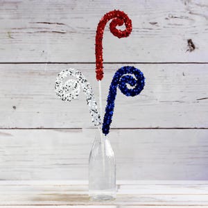 May include: Three decorative curly stems in red, white, and blue colors are arranged in a clear glass vase. The stems are made of sparkly material and resemble fireworks.