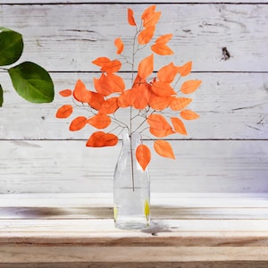 May include: A clear glass bottle vase with a bunch of orange leaves in it. The vase is sitting on a light wood surface.