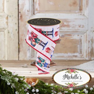 Op de afbeelding: Een rol decoratief lint met een herhalend patroon van speelgoed notenkraker soldaten in rode, groene en blauwe uniformen op een witte achtergrond. Het lint heeft rode randen en wordt weergegeven op een houten standaard. Het lint is gelabeld "D'LUX RIBBON".