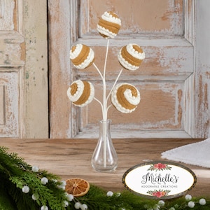 May include: A decorative arrangement featuring five tan, spherical ornaments with white spiral icing patterns, attached to white stems and displayed in a clear glass vase. An oval label with the text Michelle's Adorable Creations is visible.