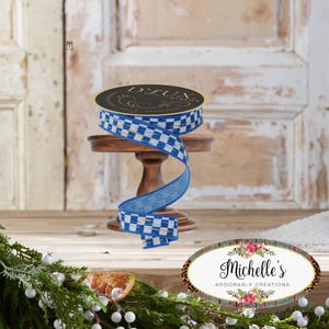 May include: A spool of D.Lux ribbon with a blue and white checkered pattern, displayed on a small wooden stand. The ribbon is partially unrolled, revealing a solid blue edge. The background includes a wooden door and decorative greenery.