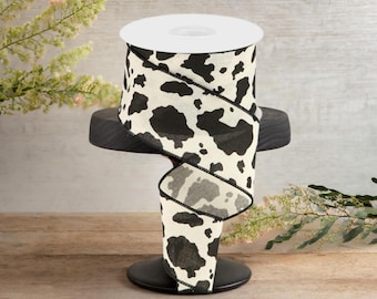 May include: A spool of white and black cow print ribbon. The ribbon is on a black stand and is ready to be used for crafting or decorating.