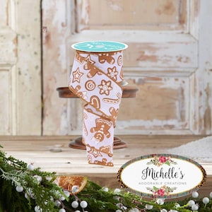May include: A spool of white ribbon featuring a repeating pattern of brown gingerbread men, candy canes, snowflakes, and swirl cookies. The ribbon has brown finished edges and is displayed on a small wooden stand. The top label reads PRG RIBBON.