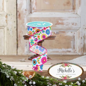 Pode incluir: Um rolo de fita floral com fundo branco e flores coloridas em tons de azul, amarelo, laranja e rosa. A fita tem uma borda rosa brilhante e é exibida em um pequeno suporte de madeira. O texto na fita diz "MG Ribbon".