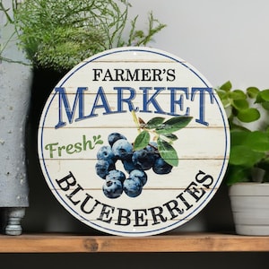 May include: A round wooden sign with whitewashed planks and blue lettering. The sign reads "Farmer's Market Fresh Blueberries". The sign is decorated with a cluster of blueberries and green leaves.
