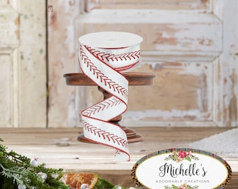 May include: A roll of white ribbon with a red stitched leaf design, displayed on a small wooden stand. The ribbon is spiraled, showcasing the pattern. The background features a rustic wooden door and decorative greenery.