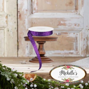 Peut inclure: Un rouleau de ruban à paillettes violet est posé sur un petit socle en bois foncé. Le ruban est déroulé, avec une partie qui pend. L'arrière-plan présente une porte rustique et des décorations végétales. Le logo de Michelle's Adorable Creations est visible.