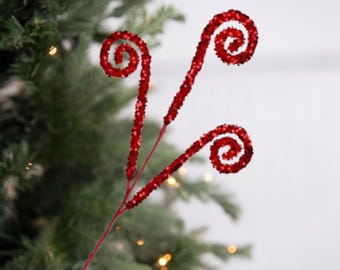 May include: Two red glittered decorative stems with a swirled design. The stems are a vibrant red colour and are placed in front of a blurred Christmas tree background.