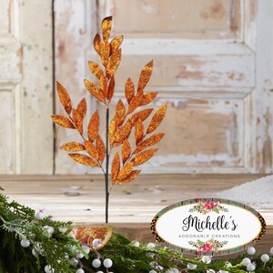 May include: A decorative orange glitter leaf stem with a black stem. The leaves are a vibrant orange with glitter, perfect for fall or holiday decor. The image also includes a sign that says "Michelle's Adorable Creations".