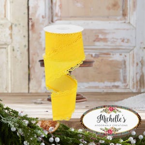 May include: A roll of bright yellow lace ribbon with a scalloped edge. The ribbon is displayed on a small wooden stand. The background features a rustic wooden door and decorative greenery. The ribbon is approximately 3 inches wide.