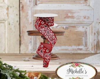 Puede incluir: Un carrete de cinta roja con un estampado de cachemira blanco, exhibido en un pequeño soporte de madera. La cinta está desenrollada, mostrando su diseño. La imagen también incluye un letrero decorativo que dice "Michelle's Adorable Creations."