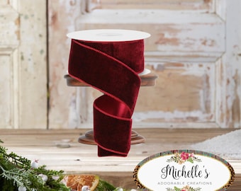 May include: A spool of burgundy velvet ribbon with a stitched edge, displayed on a small wooden stand. The ribbon is partially unrolled, revealing its texture and color. The background features a rustic wooden door and decorative greenery.