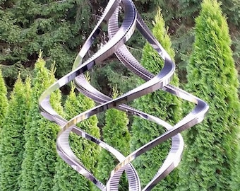 May include: A silver metal wind spinner with a geometric design. The spinner is suspended from a chain and is hanging in front of a row of evergreen trees.