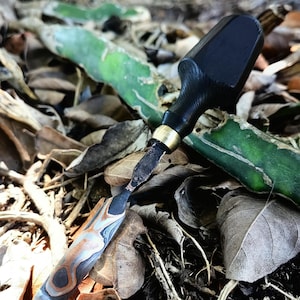 May include: A close-up of a small, hand-crafted tool with a black wooden handle and a metal blade. The blade has a unique, marbled pattern in shades of blue, brown, and gray. A brass collar connects the handle to the blade. The tool rests among dried leaves.