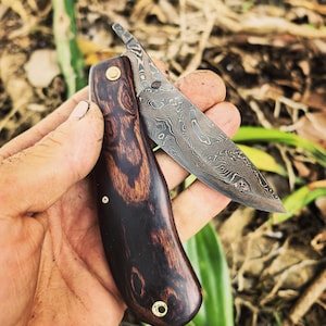 May include: A folding pocket knife featuring a dark brown wood handle with visible grain patterns and a brass pivot pin. The blade displays a distinct wavy Damascus steel pattern with a metallic grey finish and a small thumb lever for opening.