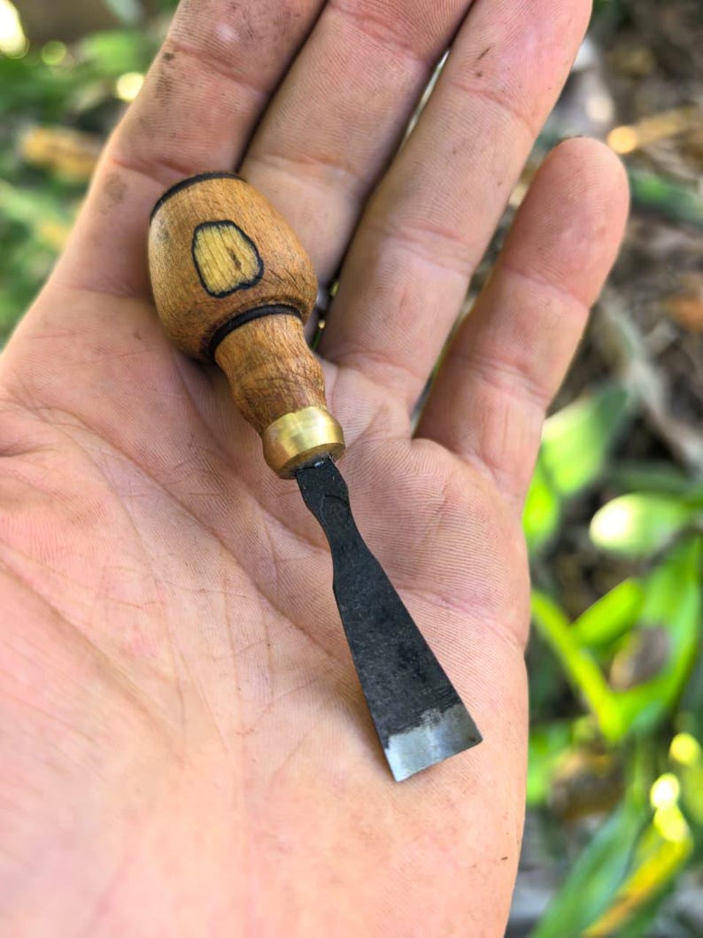 May include: A small wood chisel. It has a rounded wooden handle, brass ferrule, and a dark metal blade with a sharp edge. The handle features a light wood grain and a darker wood inlay. The chisel is approximately 15 cm long.
