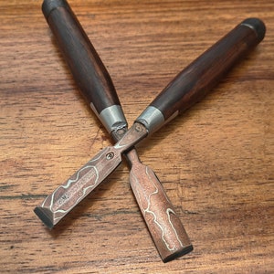 May include: Two crossed wood chisels with dark brown handles and patterned copper blades. The blades have a unique design with light and dark swirls. The chisels are set against a wooden surface.