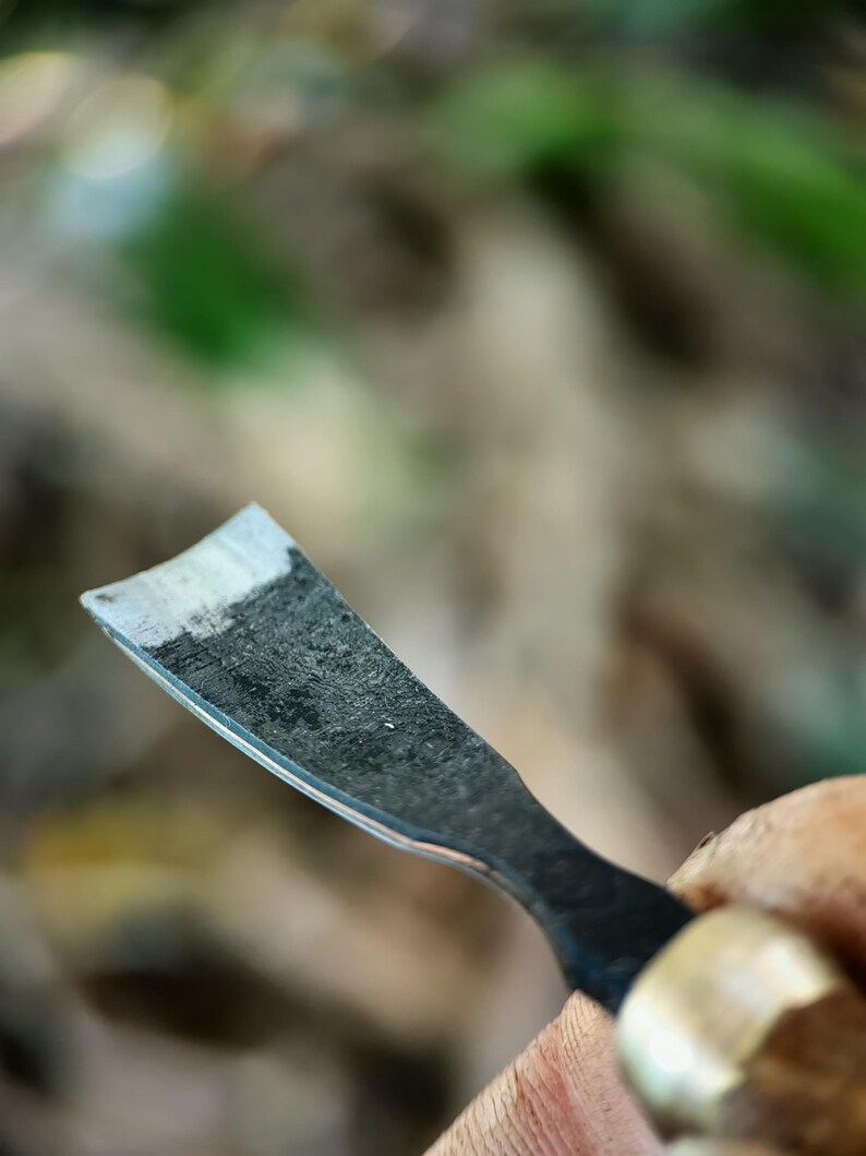 May include: Close-up of a woodworking chisel with a curved blade. The metal blade has a dark, textured appearance with a lighter, worn edge. The handle is partially visible, featuring a brass ferrule and a wooden grip.