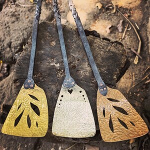 May include: Three handcrafted metal spatulas with long handles and decorative cutouts. The spatula heads are gold, silver, and bronze, with leaf and heart designs. The handles have a twisted design and a loop at the end.