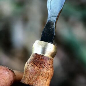 May include: Close-up of a wood-handled carving knife. The handle is light brown with a brass ferrule. The blade is silver with a patterned design. The knife is held in a hand, with a blurred background of green and brown.
