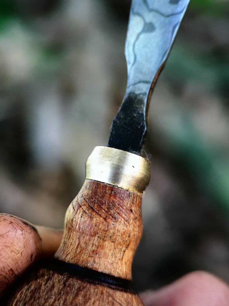 May include: Close-up of a wood-handled carving knife. The handle is light brown with a brass ferrule. The blade is silver with a patterned design. The knife is held in a hand, with a blurred background of green and brown.