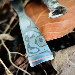 May include: A close-up shot of a metal tool with a patterned design and a copper-coloured stripe near the edge. The tool has a curved shape and is set against a backdrop of dried leaves and twigs.
