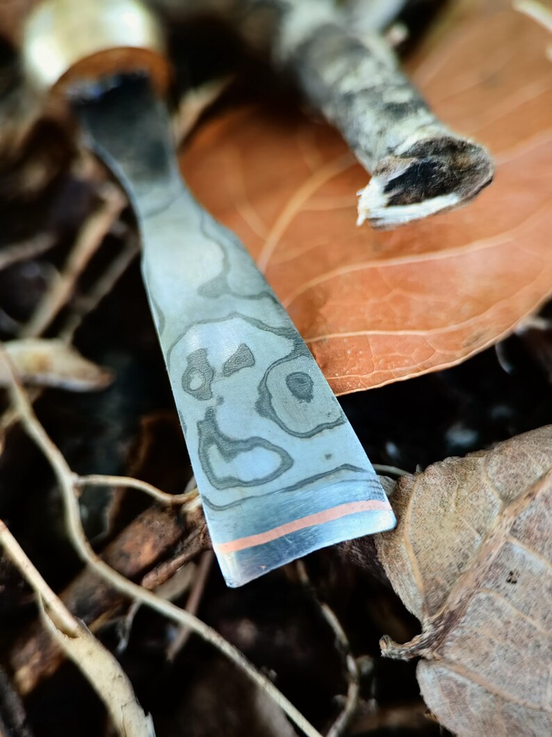 May include: A close-up shot of a metal tool with a patterned design and a copper-coloured stripe near the edge. The tool has a curved shape and is set against a backdrop of dried leaves and twigs.