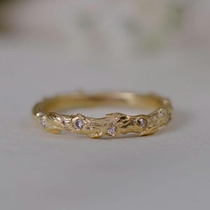 Op de afbeelding: Een gouden ring met een textuur, organisch ontwerp. De ring heeft kleine, sprankelende edelstenen langs de band. Het ontwerp van de ring lijkt op een delicate tak of wijnstok, met ingewikkelde details en een warme, gouden tint.