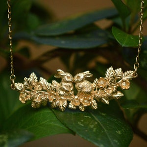 May include: A gold-toned necklace with a design of two rabbits surrounded by flowers and leaves. The necklace hangs from a fine chain. The background shows blurred green leaves. The text "by LUCY STOPES-ROE" and website address are visible.