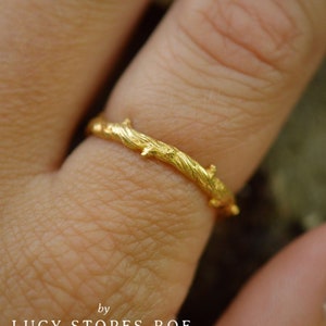 May include: A gold-colored ring with a textured twig design. The ring is worn on a finger, showcasing the intricate details of the twig-like pattern. The ring's design is inspired by nature.