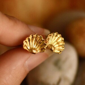 May include: A pair of gold-colored seashell stud earrings. The earrings are held between fingers and are in sharp focus. The earrings have a detailed, textured design. The text "by LUCY STOPES-ROE" and website address are visible in the background.