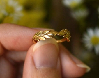 Può includere: Un anello d'oro con un delicato designo a foglia. L'anello è tenuto da una mano su uno sfondo di fiori gialli e bianchi.