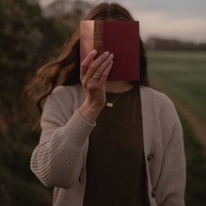 Pu&ograve; includere: Una persona tiene un libro rosso davanti al viso. Il libro ha lettere dorate sul dorso che recitano "The Vicar of Wakefield."