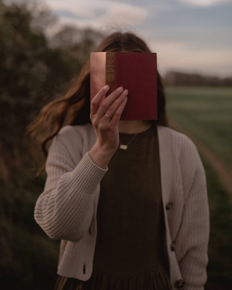 Pu&ograve; includere: Una persona tiene un libro rosso davanti al viso. Il libro ha lettere dorate sul dorso che recitano "The Vicar of Wakefield."