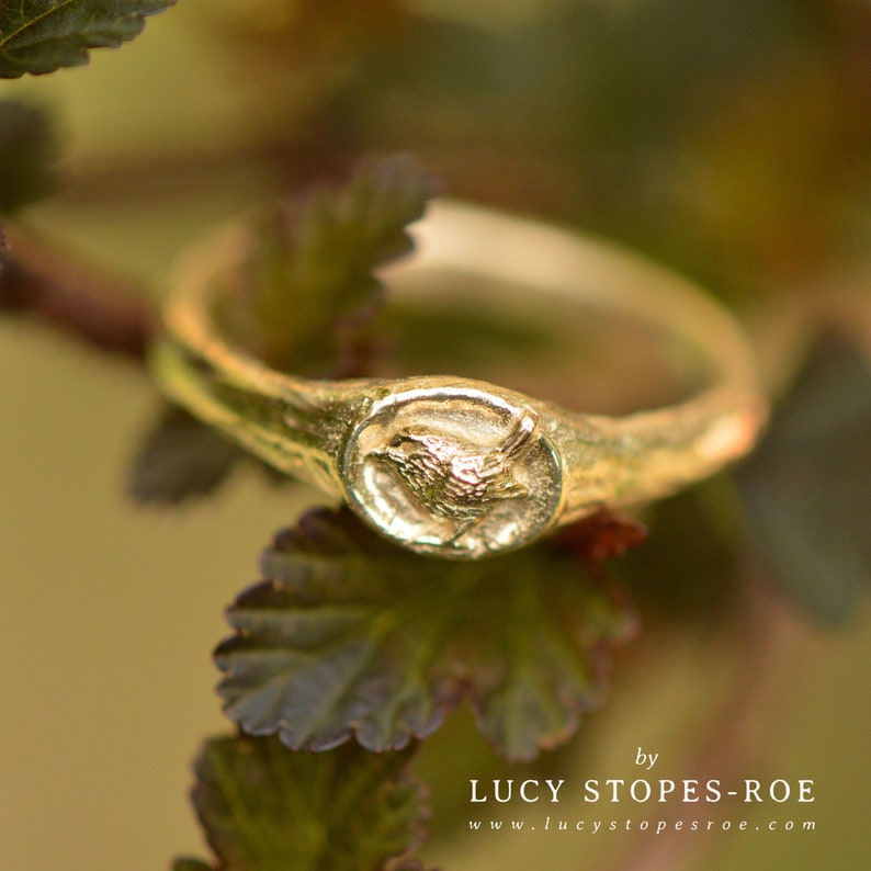 May include: A gold ring with a detailed bird design. The ring is set on a green leafy background.