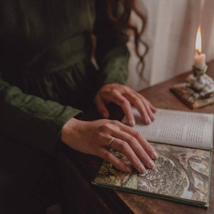 Pu&ograve; includere: Un paio di mani tengono aperto un libro con una copertina verde e illustrazioni. Il libro &egrave; su un tavolo di legno con una candela accesa sullo sfondo.