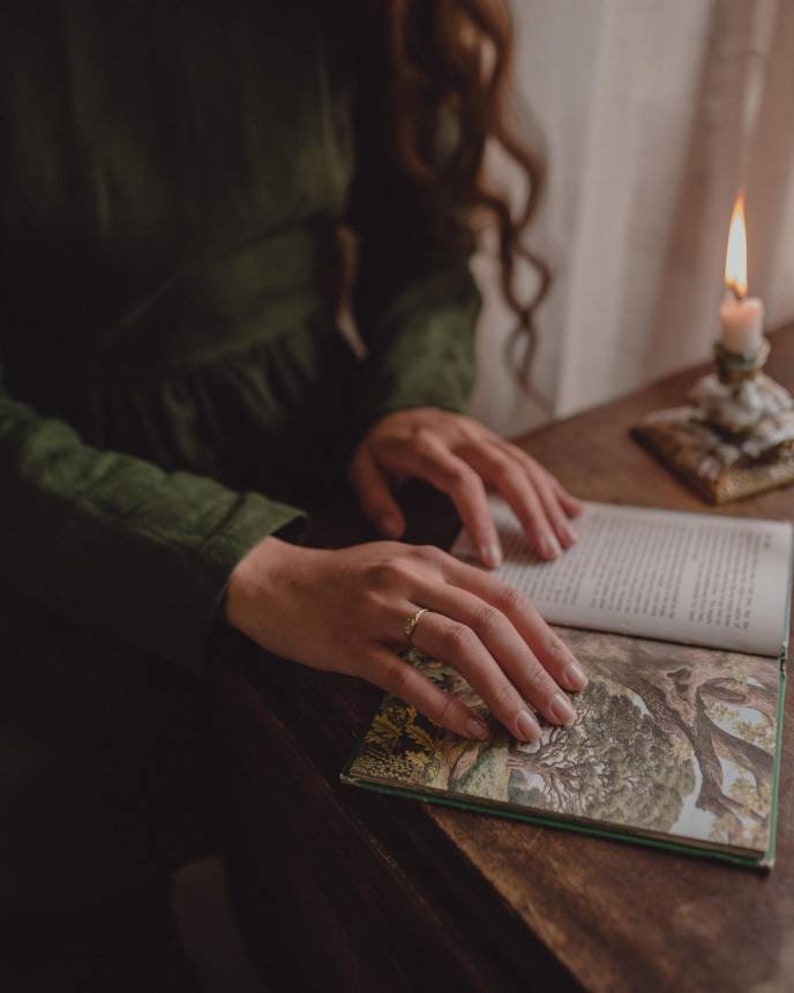 Pu&ograve; includere: Un paio di mani tengono aperto un libro con una copertina verde e illustrazioni. Il libro &egrave; su un tavolo di legno con una candela accesa sullo sfondo.