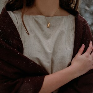 May include: A woman wearing a beige linen dress with a brown shawl draped over her shoulders. She is wearing a gold necklace with an owl pendant.