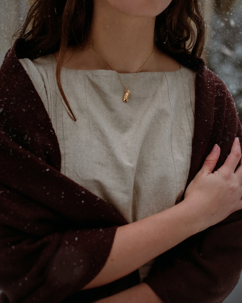 May include: A woman wearing a beige linen dress with a brown shawl draped over her shoulders. She is wearing a gold necklace with an owl pendant.