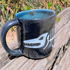 May include: Black ceramic mug with a white dinosaur skeleton design. The mug has a dark blue interior and a curved handle. The mug is sitting on a wooden surface.
