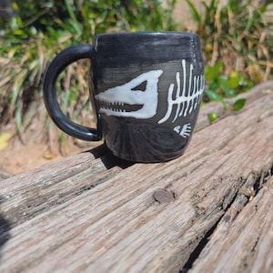 May include: Black ceramic mug featuring a white dinosaur skeleton design. The mug has a rounded shape with a black handle. The dinosaur skeleton design includes the skull and rib cage. The mug is sitting on a weathered wooden surface.