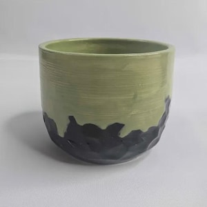 May include: A handmade ceramic bowl with a textured, two-tone design. The upper portion is a light green, while the lower half features a dark, textured, black pattern. The bowl has a cylindrical shape and a smooth interior.