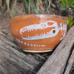 May include: A handmade ceramic bowl with a brown exterior and a white dinosaur skeleton design. The bowl is round with a slightly flared rim. The dinosaur skeleton design is a prominent feature, adding a unique touch to the bowl's aesthetic.