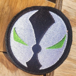May include: A white and black embroidered patch with a green symbol. The symbol is a stylized face with two green eyes and a black mouth.