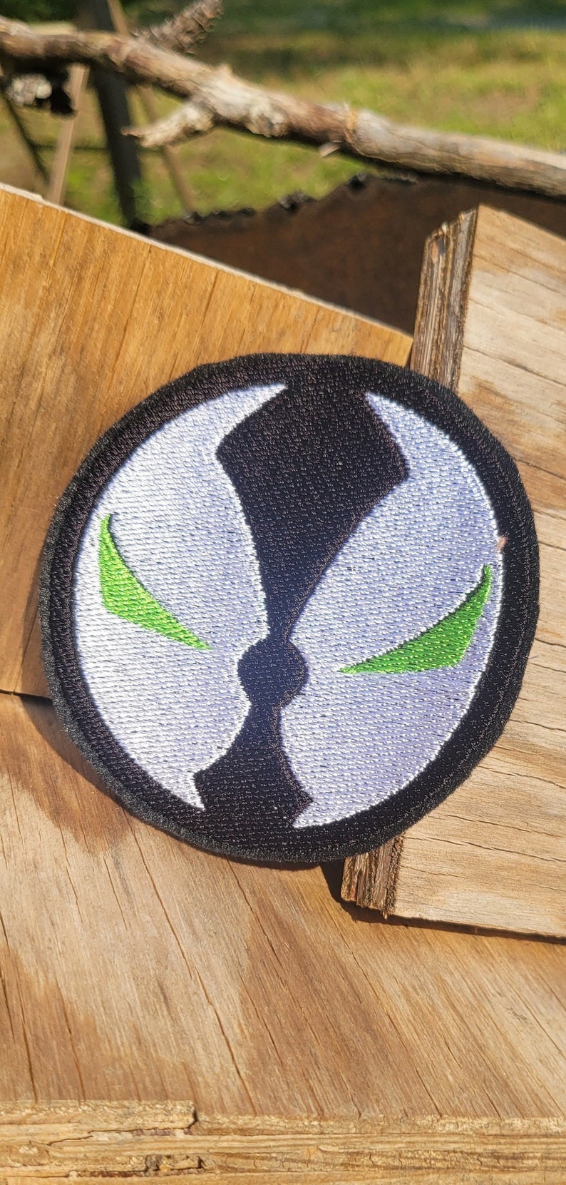 May include: A white and black embroidered patch with a green symbol. The symbol is a stylized face with two green eyes and a black mouth.