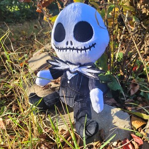 Op de afbeelding: Een pluche speelgoed van Jack Skellington uit de film Nightmare Before Christmas. Het speelgoed zit op een rots met herfstbladeren en gras eromheen. Het speelgoed draagt een zwart-wit gestreept pak met een witte kraag en een zwarte strik.